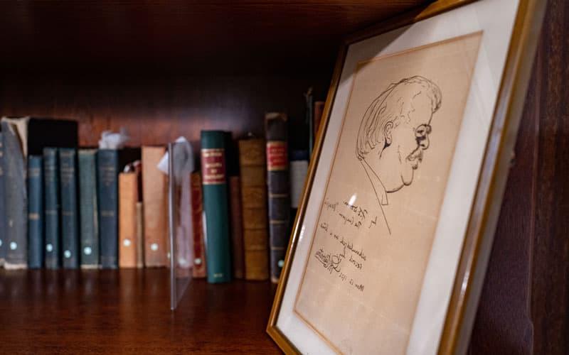A caricature of GK Chesterton in a frame, sitting to next to a shelf with all his books.