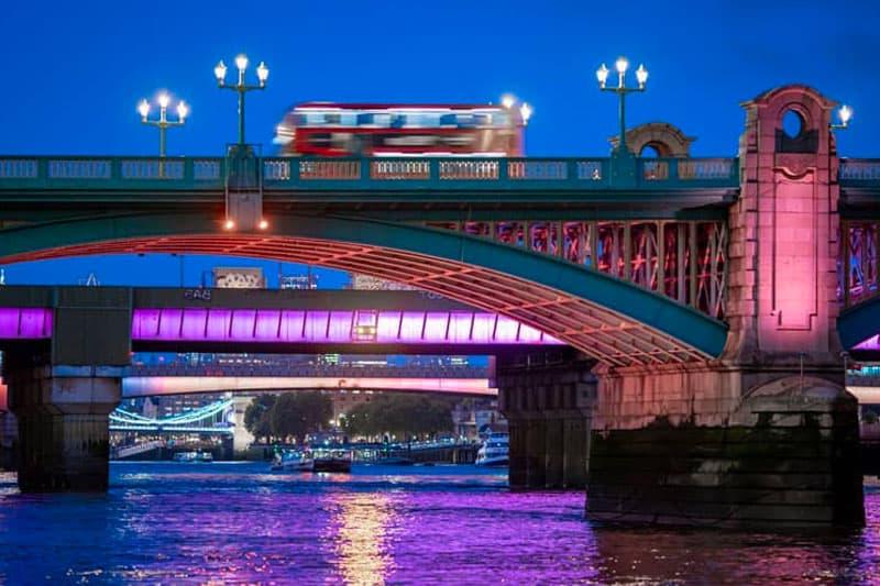 A image of a blurry red double-decker buse as it travels on a bridge over the River Thames at night.